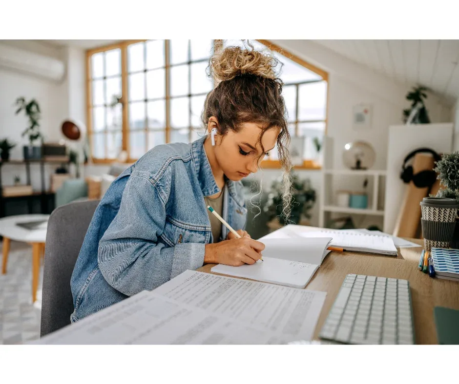 Como superar os desafios do ensino superior à distância? | Foto de uma jovem com fone de ouvido sentada em uma mesa com o notebook e fazendo anotações em um caderno | UNIFAVENI