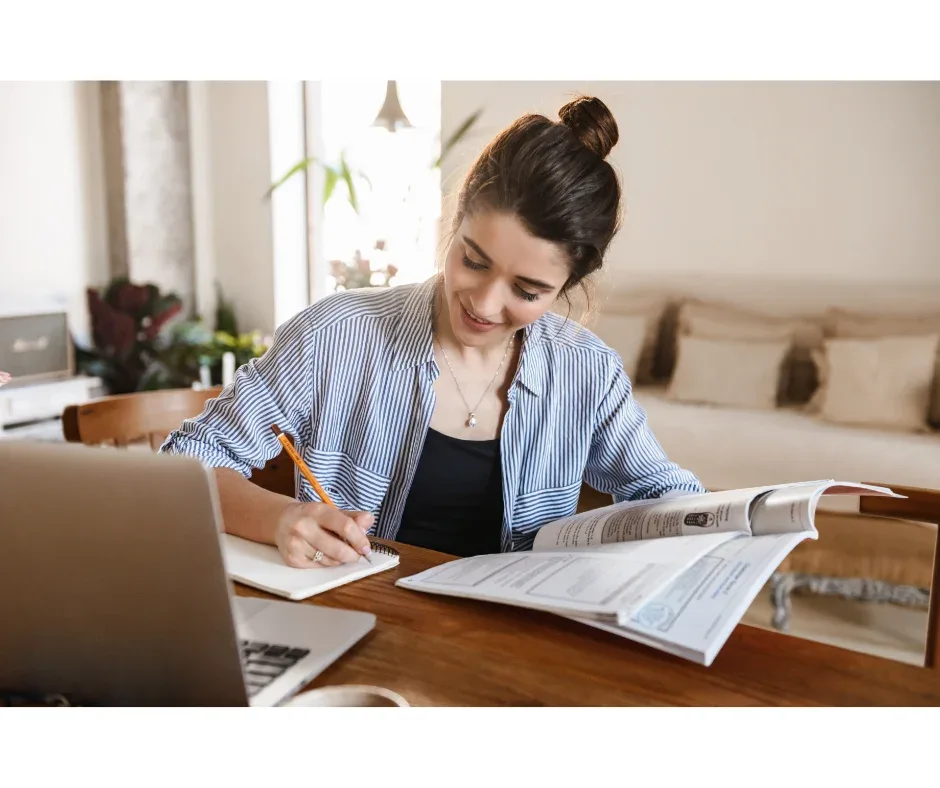 Custos na graduação EaD e na presencial: qual a diferença? | Jovem mulher lendo um livro e fazendo anotações em frente ao notebook | UNIFAVENI