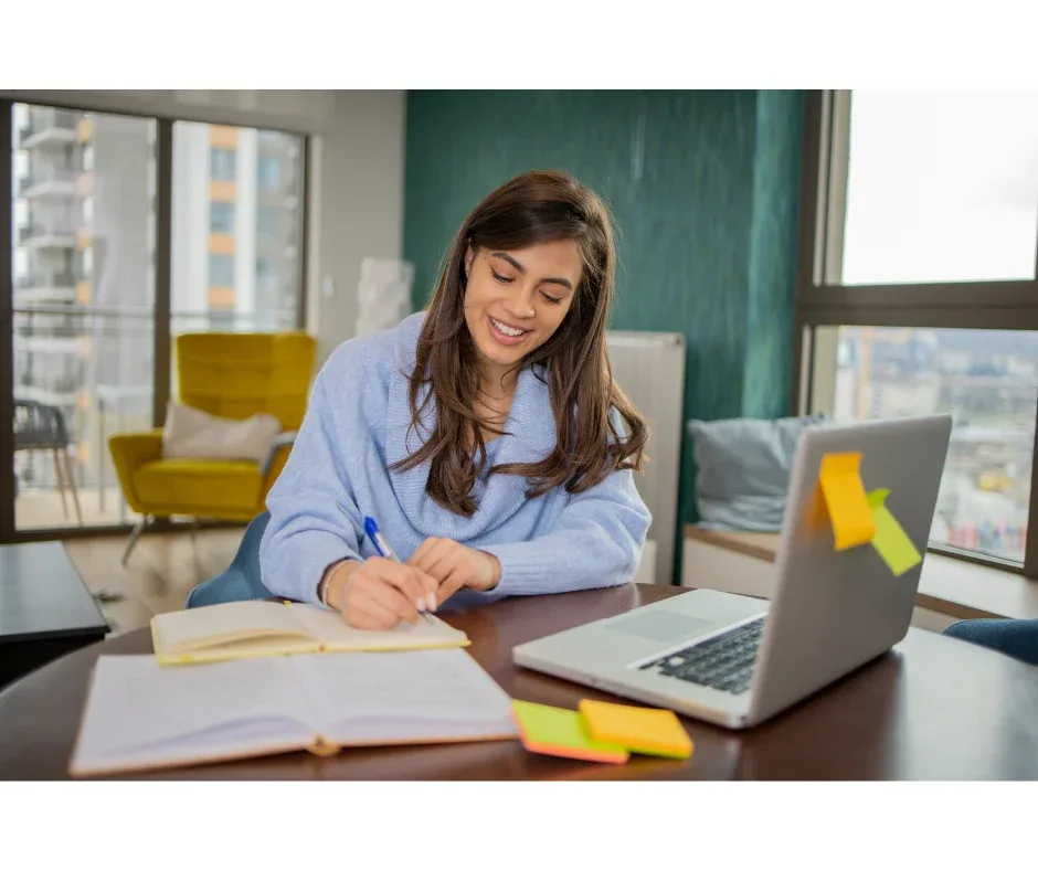 Estudos na graduação EaD: como conciliar com a rotina? | Jovem mulher sorrindo enquanto faz anotações na frente do notebook | UNIFAVENI