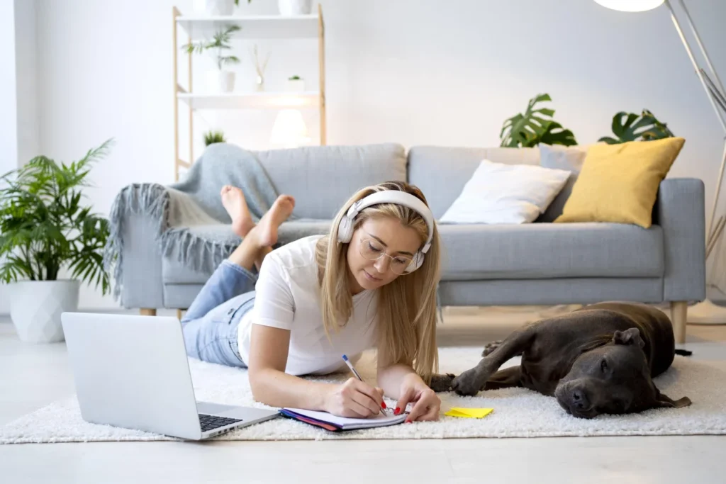 Graduação EaD para deficientes visuais: principais vantagens | Jovem mulher deitada no chão da sala mexendo no notebook e um cachorro ao lado | UNIFAVENI