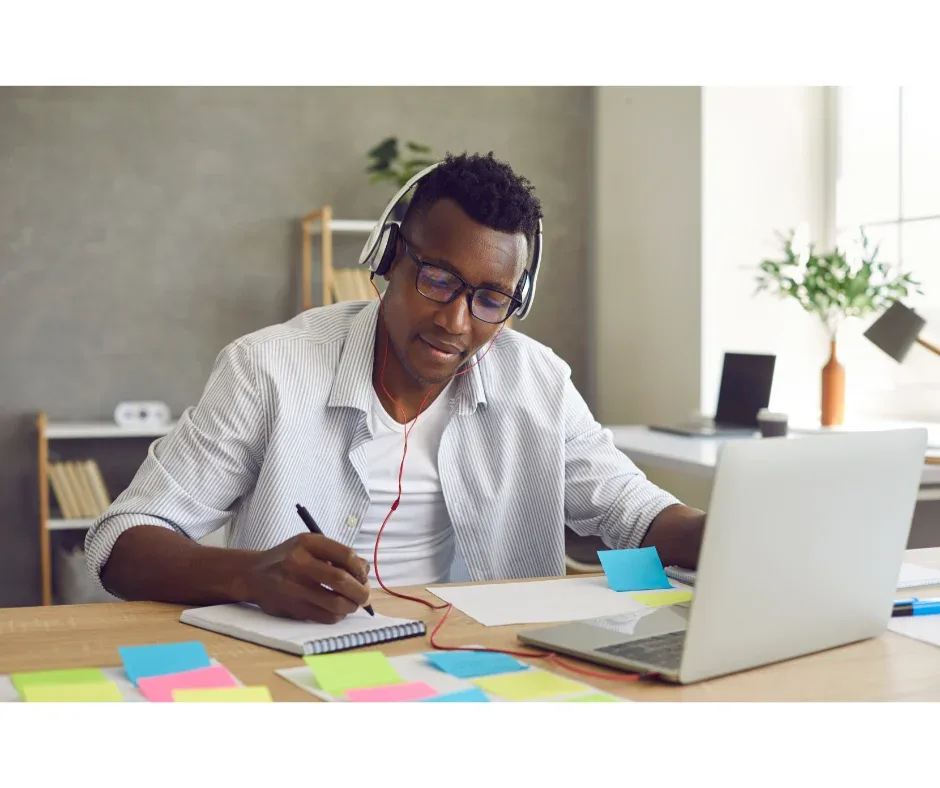 Sucesso acadêmico em EaD: quais são os recursos disponíveis? | Jovem rapaz com fones de ouvido fazendo anotações em um caderno em frente ao notebook  | UniFaveni