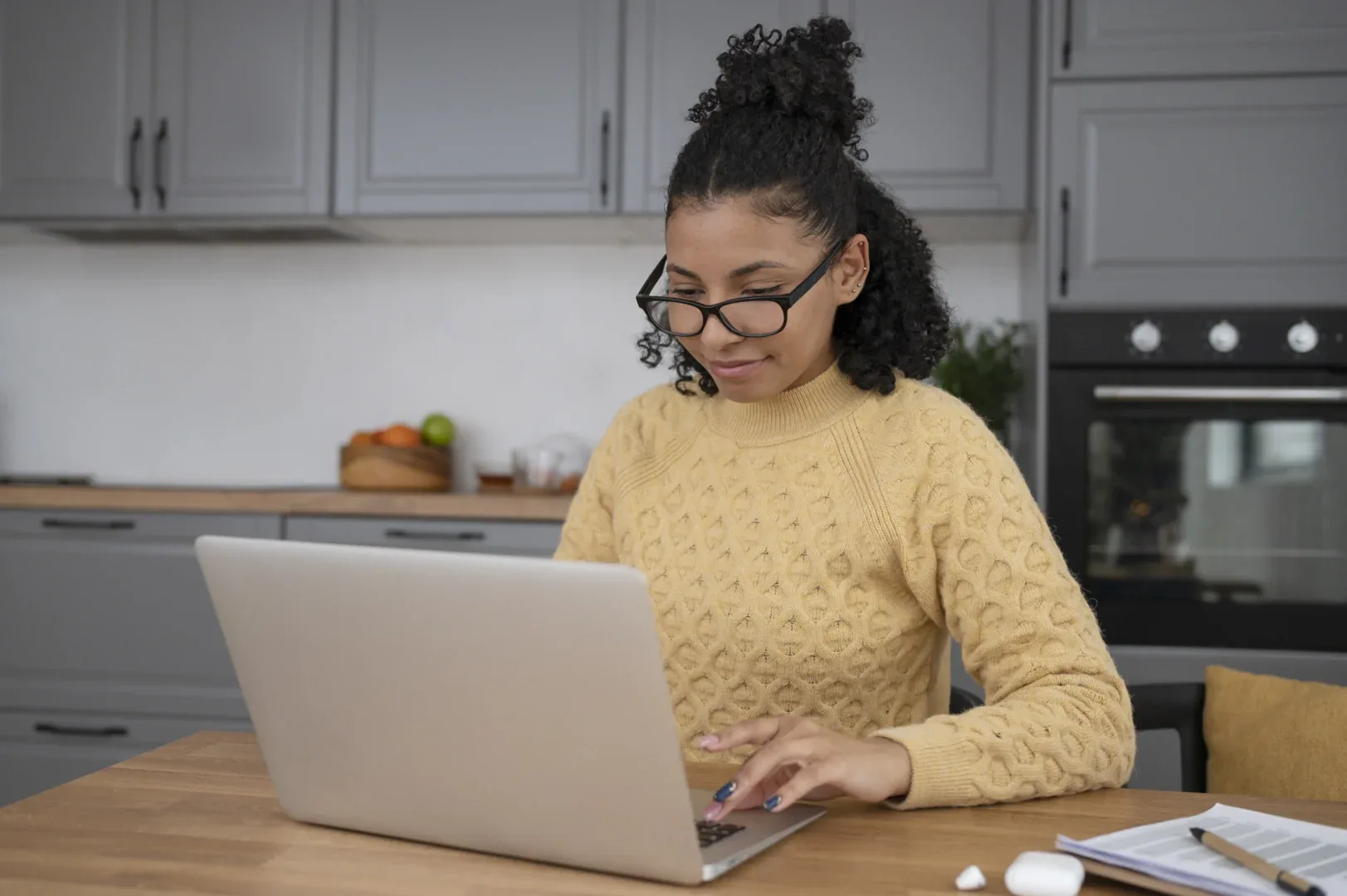 O TCC na graduação EaD é obrigatório? | Foto de jovem sentada na bancada da cozinha usando óculos e mexendo no notebook | UNIFAVENI