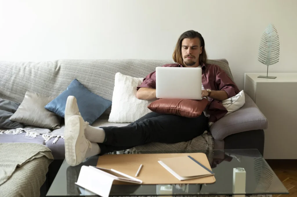 Atividades complementares na graduação EaD: como conseguir? | Jovem homem sentado no sofá mexendo no notebook com uma expressão séria | UNIFAVENI|Atividades complementares na graduação EaD: como conseguir? | Jovem homem sentado no sofá mexendo no notebook com uma expressão séria | UNIFAVENI